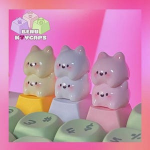 May include: Three sets of cat-shaped keycaps in cream, light gray, and pink, stacked on colorful keyboard keys. The keycaps have black eyes and rosy cheeks. The brand name "BERU KEYCAPS" is displayed in a logo at the top.