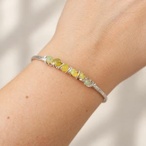 Puede incluir: Un brazalete de metal plateado que presenta una fila de seis pequeñas cuentas redondas e iridiscentes similares a ópalos. Las cuentas están aseguradas al centro de la banda de metal con textura martillada mediante un fino alambre plateado.