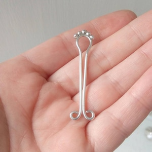 Puede incluir: Una pinza para pezones plateada con un diseño floral en la parte superior y un diseño en bucle en la parte inferior. La pinza se sostiene en una mano sobre un fondo neutro.