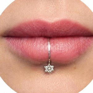 Könnte beinhalten: Nahaufnahme von Lippen mit einem silbernen Lippenring. Der Lippenring ist ein kleiner Reifen mit einem anhängenden blütenförmigen Anhänger. Die Lippen haben eine natürliche rosa Farbe.