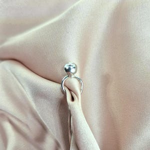 Op de afbeelding: Een zilveren sjaalring met een kleine bal aan de bovenkant en een cirkelvormig ontwerp. De ring staat tegen een achtergrond van licht perzik satijnen stof, wat een zachte, elegante uitstraling creëert.