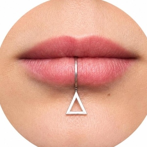 Puede incluir: Primer plano de unos labios con un piercing labial plateado. El piercing es un pequeño aro con un colgante triangular. La piel alrededor de los labios es lisa y los labios son de color rosa natural.