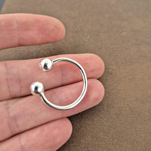 May include: A silver-colored open ring featuring a smooth, polished band that terminates at both ends with a small, spherical ball. The metal has a reflective, metallic finish, and the design is circular with a gap between the two spheres.