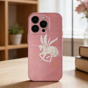 May include: A pink phone case featuring a white winged unicorn design. The case has a glossy finish and a camera module with multiple lenses. The phone case is on a wooden table with a blurred background.