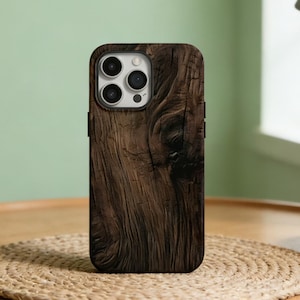 May include: A brown wooden phone case with a textured wood grain design. The case has a camera cutout and is displayed on a round wooden table with a woven placemat. A potted plant and a lamp are in the background.