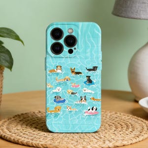 May include: A turquoise phone case featuring a pattern of dogs swimming in a pool. The case has a textured water design and a collection of various dog breeds floating in the water. The case is on a wooden table with a woven placemat.