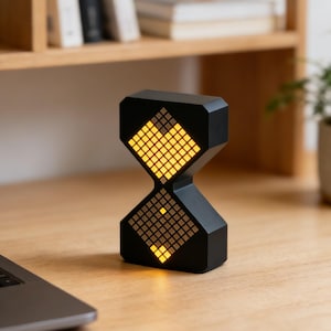 May include: A black hourglass-shaped digital clock with a yellow illuminated display. The clock is on a wooden desk next to a laptop and a small potted plant. The clock's display is made up of small square pixels.