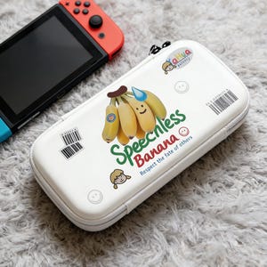 May include: A Nintendo Switch console with red and blue controllers next to a white case. The case features a banana graphic with the text "Speechless Banana" and a barcode sticker.