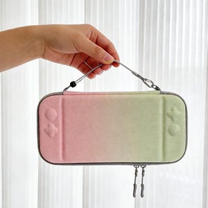 Puede incluir: Una funda de transporte con degradado rosa y verde para una consola de juegos. La funda tiene un ribete gris, un cierre de cremallera y un asa plateada. La funda está sujeta por una mano.