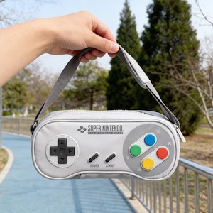 Pode incluir: Uma bolsa cinza em forma de controle Super Nintendo com uma alça cinza. A bolsa possui fecho de zíper e botões coloridos. O texto "SUPER NINTENDO" está impresso na bolsa.