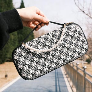 May include: A black and white plaid pattern handbag with a pearl strap. The rectangular bag has a black handle and a gold zipper. The bag is held up by a person's hand.