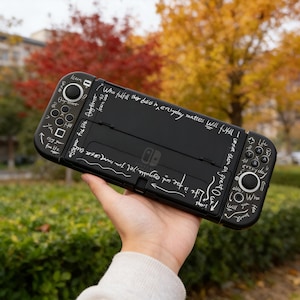 May include: Black Nintendo Switch console with white handwritten text. The text includes phrases like "Who fulfill their duties in everyday matters will fulfill" and "Enjoy your life". The console has black buttons and joysticks.