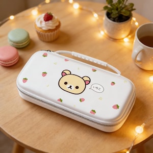 Puede incluir: Estuche rectangular blanco con una ilustración de oso de dibujos animados, fresas y texto japonés. El estuche tiene un asa y cierre de cremallera. Otros elementos en la mesa incluyen una magdalena, macarons y una taza de té.