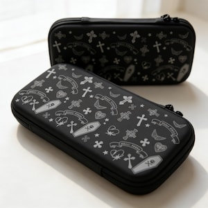 Op de afbeelding: Twee zwarte etuis met ritssluiting met een herhalend patroon van kruisen, doodskisten, harten, vleugels en banners. De banners lezen "MAGIC KISSES" in een gotisch lettertype. De etuis zijn rechthoekig en hebben een gladde textuur.