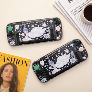 Op de afbeelding: Twee zwarte Nintendo Switch console cases met een fantasieontwerp. Elke case heeft een engel illustratie met witte vleugels, omringd door sterren, harten en andere decoratieve elementen. De cases hebben kleurrijke knoppen.