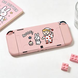 May include: A pink Nintendo Switch console with cartoon stickers, including a bunny, a boy in a polka-dot outfit, and the word "NICE!". A pink notebook with the words "BE BETTER" is visible in the background. Two pink building blocks are also present.
