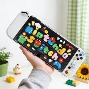 Op de afbeelding: Een wit en zwarte Nintendo Switch-console met een doorzichtige beschermhoes. De hoes is versierd met kleurrijke cartoonafbeeldingen en het woord "Nintendo Switch". De console wordt in de hand van een persoon gehouden.