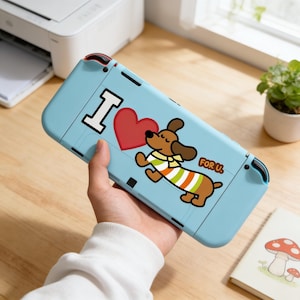 Op de afbeelding: Een lichtblauwe Nintendo Switch-hoes met een cartoon teckel die een rood hart vasthoudt. De hoes heeft de tekst "I" en "FOR U.". De teckel draagt een gestreept shirt met groene, oranje en witte strepen.