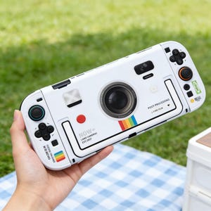 May include: White Nintendo Switch case designed to resemble a vintage camera. The case features a large lens, a red button, and a rainbow stripe. The text "NOW+ LAND CAMERAS EST.1937" is printed on the case.