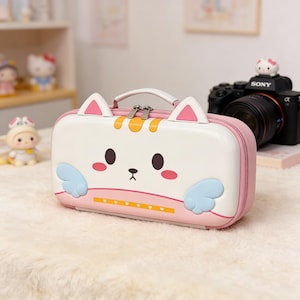 May include: A white and pink cat-themed travel case with a handle and zipper closure. The case features a cartoon cat face with pink cheeks, brown eyes, and blue wings. The case is on a fluffy white surface.