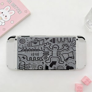 Op de afbeelding: Een grijze Nintendo Switch-console met een doodle-ontwerp. Het ontwerp bevat een stokfiguur, enveloppen en andere vormen. De console heeft witte controllers en een roze notitieboek met de tekst "BE BETTER".