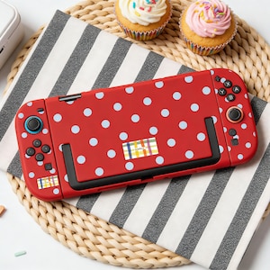 以下が含まれることがあります： 赤色のNintendo Switchコンソール。水色のドット模様。コンソールには黒いボタンと小さなカラフルなステッカーが付いています。背景には、フロスティングとスプリンクルが施された2つのカップケーキがあります。