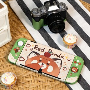 May include: A Nintendo Switch console with a custom Red Panda design, featuring a cartoon red panda with an apple on its head. The console is green and white, with the text "Red Panda". A Fujifilm X-T5 camera and cupcakes are also present.