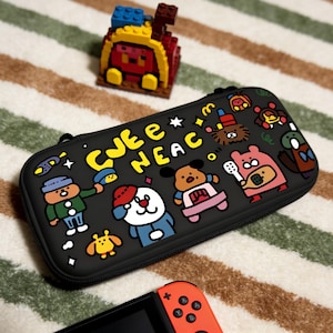 May include: Black carrying case with colorful cartoon characters and the text "CJEE NEAC". A small Lego structure is in the background. An orange and black game controller is in the foreground.