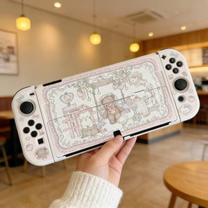Pode incluir: Uma capa branca e rosa para Nintendo Switch com uma ilustração fantasiosa de ursinhos de pelúcia, gatos e elementos florais. A capa tem uma borda decorativa e é segurada na mão de uma pessoa.