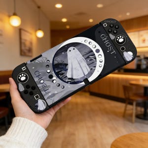 Könnte beinhalten: Schwarze und graue Nintendo Switch-Hülle mit Geisterdesign. Die Hülle hat eine weiße Geisterillustration in einer Mondform, mit dem Wort "GHOST" auf der Seite. Sie hat auch Pfotenabdruck- und Geister-Icons.