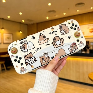 Puede incluir: Una funda de consola de juegos de color crema con ilustraciones de osos de dibujos animados. La funda tiene botones negros y un joystick con un diseño de cara sonriente. El texto incluye "Haha" y "abc".