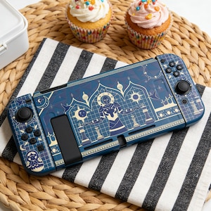 May include: A blue and gold patterned Nintendo Switch console sits on a black and white striped cloth. The console features a detailed design with figures and geometric shapes. Two cupcakes with frosting and sprinkles are in the background.