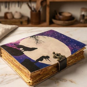 Könnte beinhalten: Ein handgefertigtes Notizbuch mit einem Himmelsdesign mit Wölfen unter einem Vollmond. Das Cover hat einen lila und blauen Sternenhimmel mit einem großen weißen Mond. Das Notizbuch ist mit einem schwarzen Lederriemen und einem Metallverschluss gesichert.