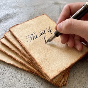 May include: A stack of rectangular, textured, beige paper sheets with deckled edges. A black fountain pen is writing the words The art of on the top sheet. The paper has a fibrous, handmade appearance with darkened, uneven edges.
