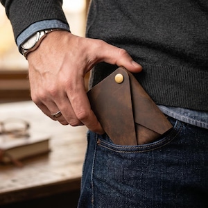 May include: A dark brown leather card holder with an envelope-style flap and a single gold-toned metal snap button closure. The card holder is rectangular in shape and is being placed into the front pocket of a pair of dark blue denim jeans.