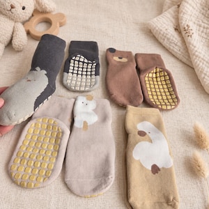 May include: A collection of six infant socks in various neutral shades including charcoal, brown, beige, and tan. Each sock features a whimsical animal graphic and a yellow or white rubberised grid pattern on the sole for added traction and grip.