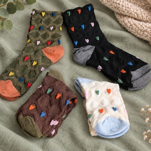 May include: Four pairs of crew socks featuring a textured diamond knit pattern and small, raised, multi-colored heart accents. The socks come in olive green with orange accents, black with grey accents, dark brown with olive accents, and white with blue accents.