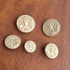 May include: Five circular gold-toned buttons of varying sizes, each featuring a raised relief design of a figure with flowing hair. The buttons have a smooth, metallic finish and a decorative border, arranged in a cluster on a textured surface.