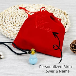 Puede incluir: Un collar con colgante azul claro con una parte superior dorada, presentado con una bolsa de terciopelo roja. La bolsa está bordada con el nombre "Becky" y un diseño floral. También es visible el texto "Personalized Birth Flower & Name".