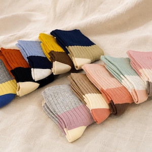 Könnte beinhalten: Eine Sammlung gerippter Socken, angeordnet in zwei Reihen, mit verschiedenen Color-Block-Designs in Grau, Orange, Blau, Gelb, Braun, Marineblau, Creme, Rosa, Grün und Beige. Jedes Paar zeigt eine eigene Kombination aus verschiedenen Farben.