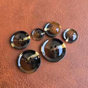 May include: A collection of six round, four-hole buttons in varying sizes. Each button features a glossy, dark brown tortoiseshell pattern with a distinct, light-colored diagonal streak across the surface, creating a smooth, reflective finish.