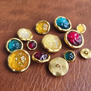 May include: A collection of round buttons featuring gold-toned metal rims and translucent, textured centers in shades of yellow, red, and teal. The buttons vary in size, with some displaying intricate patterns on the surface and others showing a ribbed metal back.
