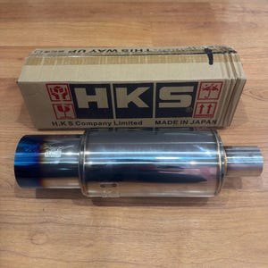 Könnte beinhalten: Ein Auspuffschalldämpfer aus Edelstahl mit blau getöntem Ende, neben einem Karton. Die Box zeigt das HKS-Logo und den Text "H.K.S Company Limited MADE IN JAPAN". Der Schalldämpfer ist zylindrisch und hat eine polierte Oberfläche.