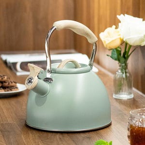 May include: A light green tea kettle with a wooden handle and spout detail. The kettle has a stainless steel frame and a wooden handle. The kettle is sitting on a wooden surface. A small vase of flowers is in the background.