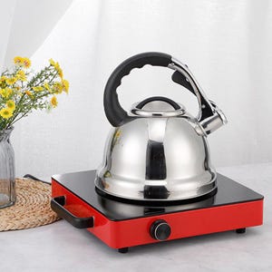 May include: A stainless steel stovetop kettle with a black handle and spout sits on a red and black electric burner. The kettle is round and shiny, reflecting the light. A vase of yellow flowers is in the background.