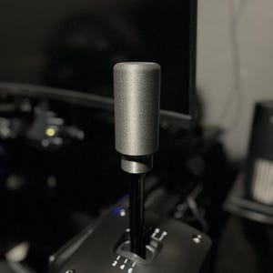 Puede incluir: Una palanca de cambios Fanatec negra y plateada con un mango cilíndrico plateado. La base de la palanca de cambios tiene marcas de engranajes y el logotipo de Fanatec. El fondo está borroso, mostrando un monitor y otros equipos.