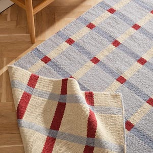 May include: A woven area rug featuring a grid pattern of light blue and cream squares intersected by bold red lines. The rug has a textured, fabric-like surface and one corner is folded over to reveal the underside, showing the same plaid design.