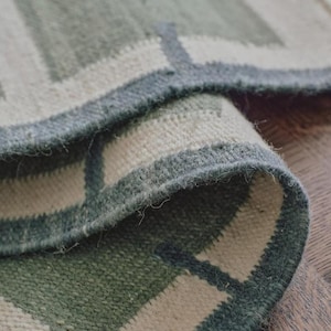 May include: A woven rug featuring a geometric pattern of sage green and off-white rectangular blocks. The fabric has a visible textured weave with a thick, dark green border outlining the shapes, showing a folded corner revealing the underside.