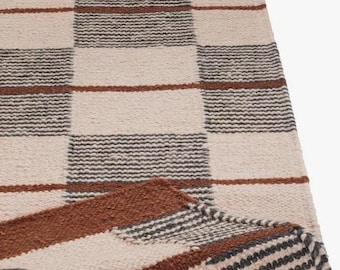 May include: A woven area rug featuring a large-scale checkered pattern in cream, charcoal gray, and rust brown. The rug has a textured, looped pile construction with a corner folded over to reveal the underside and the thickness of the weave.