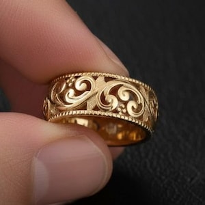 以下が含まれることがあります： 精巧な透かし彫りデザインのゴールドカラーのリング。リングは、渦巻く花のようなモチーフの繰り返し模様が特徴です。リングは暗い背景に配置され、その詳細な職人技と温かみのある色合いを際立たせています。